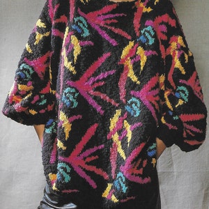 Könnte beinhalten: Ein schwarzer Pullover mit einem bunten abstrakten Muster. Das Muster zeigt rosa, gelbe, blaue und grüne geometrische Formen.