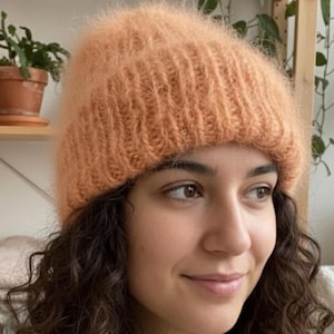 Könnte beinhalten: Eine pfirsichfarbene, gestrickte Beanie-Mütze. Die Mütze besteht aus einem weichen, flauschigen Material und hat eine gerippte Textur. Die Mütze wird auf dem Kopf getragen. Die Beanie ist ein warmes und stilvolles Accessoire für kaltes Wetter.