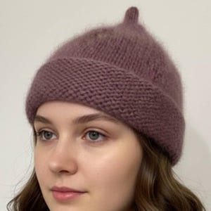 Könnte beinhalten: Eine mauvefarbene Strickmütze mit geripptem Saum und kleinem, spitzem Oberteil. Die Mütze ist aus einem weichen, flauschigen Material gefertigt und soll den Kopf warm halten. Ein stilvolles Accessoire für kaltes Wetter.