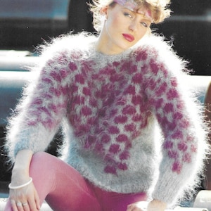 May include: A fluffy, grey and pink sweater with a leopard print design. The sweater is being worn by a person with blonde hair and a pink headband. The person is sitting on a metal surface.