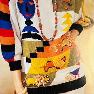 Peut inclure: Un pull en tricot blanc avec un motif géométrique coloré. Le pull présente un visage bleu, des rayures orange et jaunes, et un motif animal brun et vert. Le pull est décoré de perles brunes et d'un long collier.