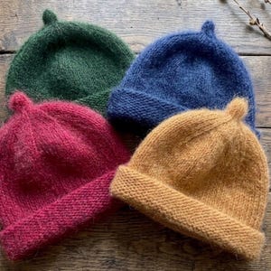 Könnte beinhalten: Vier gestrickte Mützen in Dunkelgrün, Marineblau, Magenta und Senfgelb. Jede Mütze hat eine flauschige, gebürstete Textur, einen umgeschlagenen Rand und ein kleines spitzes Detail an der Oberseite. Die Mützen sind zusammen angeordnet.