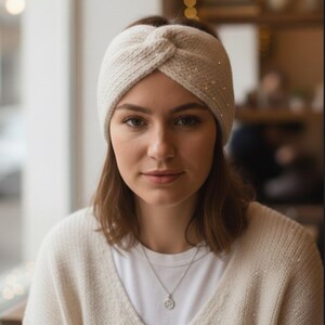 Könnte beinhalten: Ein cremefarbenes Stirnband aus Strick mit einem verdrehten Knotendesign. Das Stirnband hat kleine, glitzernde Verzierungen entlang der Oberseite. Die Person auf dem Bild trägt einen cremefarbenen Pullover und ein weißes T-Shirt.