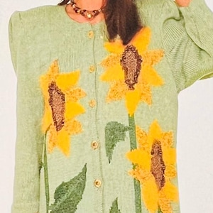 Könnte beinhalten: Ein grüner Strickcardigan mit Sonnenblumenmuster. Der Cardigan hat einen Knopfleistenverschluss und einen breiten Kragen. Das Model trägt einen passenden Hut.