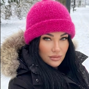 Könnte beinhalten: Eine leuchtend pinkfarbene Strickmütze mit abgerundeter Krone. Die Mütze wird mit einem schwarzen Wintermantel mit fellgefütterter Kapuze getragen. Der Hintergrund zeigt eine verschneite Winterlandschaft.