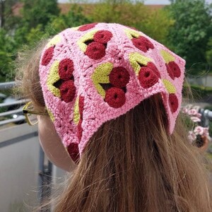 Könnte beinhalten: Ein rosa gehäkeltes Stirnband mit einem Muster aus roten und grünen Kirschen. Das Stirnband wird auf dem Kopf einer Person getragen.