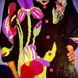 Könnte beinhalten: Schwarzer Cardigan mit einem farbenfrohen, gestickten Blumendesign. Die Blumen sind rosa, lila, gelb und grün. Der Cardigan hat einen Knopfverschluss.