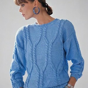 Könnte beinhalten: Hellblauer Zopfstrickpullover mit Rundhalsausschnitt und langen Ärmeln. Der Pullover hat ein strukturiertes Zopfmuster in der Mitte und an den Ärmeln. Die Person trägt blaue Creolen und einen karierten Rock.
