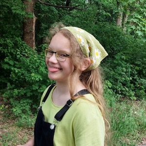 May include: A young person with long blonde hair wearing a green and white bandana with a daisy pattern, yellow-framed glasses, and a green shirt with black overalls.