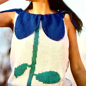 Könnte beinhalten: Ein weißes ärmelloses Strickoberteil mit einem blauen Kragen und einem grünen Stiel- und Blattdesign. Das Oberteil wird mit einem blau-grünen gestreiften Rock getragen.