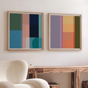 May include: Two framed abstract art prints with geometric shapes in shades of blue, green, pink, and orange. The prints hang above a wooden console table with two wooden storage boxes. A cream-coloured, rounded armchair and a woven rug complete the room.