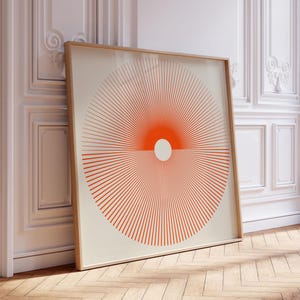 May include: A framed art print featuring a geometric design of radiating orange lines and a central white circle against a cream background. The artwork is displayed in a room with white walls and a wooden floor.