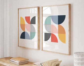 Puede incluir: Dos impresiones de arte abstracto enmarcadas con formas geométricas en tonos de azul, verde, amarillo, naranja, rojo y rosa. Las impresiones están colgadas en una pared blanca sobre una mesa consola de madera con libros y una lámpara.