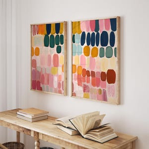 May include: Two framed abstract art prints with colorful, vertical oval shapes in shades of pink, orange, yellow, green, and blue. The artwork hangs above a wooden table with books and a wicker basket.