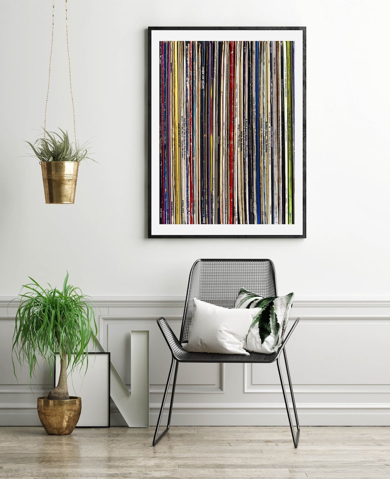 Pu&ograve; includere: Una stampa incorniciata nera di un design astratto colorato fatto di strisce verticali di vari colori e trame. La stampa &egrave; appesa a un muro bianco sopra una sedia in metallo nero con un cuscino bianco.