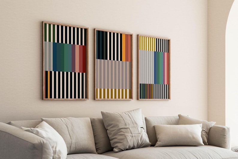 Pode incluir: Tr&ecirc;s impress&otilde;es de arte abstrata emolduradas com listras verticais coloridas em uma sala de estar. As impress&otilde;es est&atilde;o penduradas em uma parede branca acima de um sof&aacute; cinza claro com almofadas.