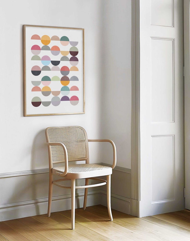 May include: A framed abstract print with a geometric pattern of colorful semi-circles in various shades of red, orange, yellow, green, blue, purple, and gray. The print is hanging on a white wall next to a light wood chair with a woven cane seat.