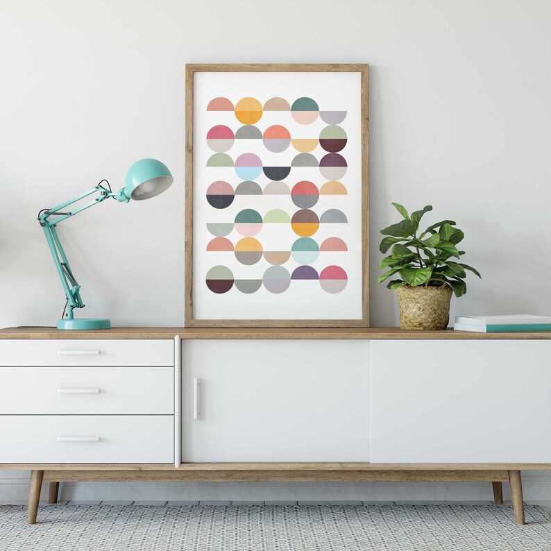 May include: A white dresser with three drawers and two cabinet doors. A framed print with a geometric pattern of colorful semi-circles hangs above the dresser. A turquoise desk lamp and a potted plant are also on the dresser.