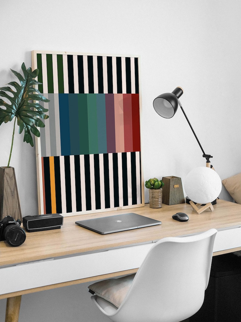Pode incluir: Uma impress&atilde;o de arte abstrata emoldurada com listras verticais pretas e brancas e uma fileira de listras coloridas no meio. A impress&atilde;o est&aacute; pendurada em uma parede branca acima de uma mesa com um laptop, uma l&acirc;mpada e uma planta em vaso.