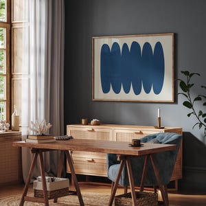 May include: A dark blue abstract art print with a white background hangs on a dark gray wall above a wooden dresser with woven cane drawers. A blue armchair sits in front of the dresser, and a wooden table with a trestle base sits in front of the chair.