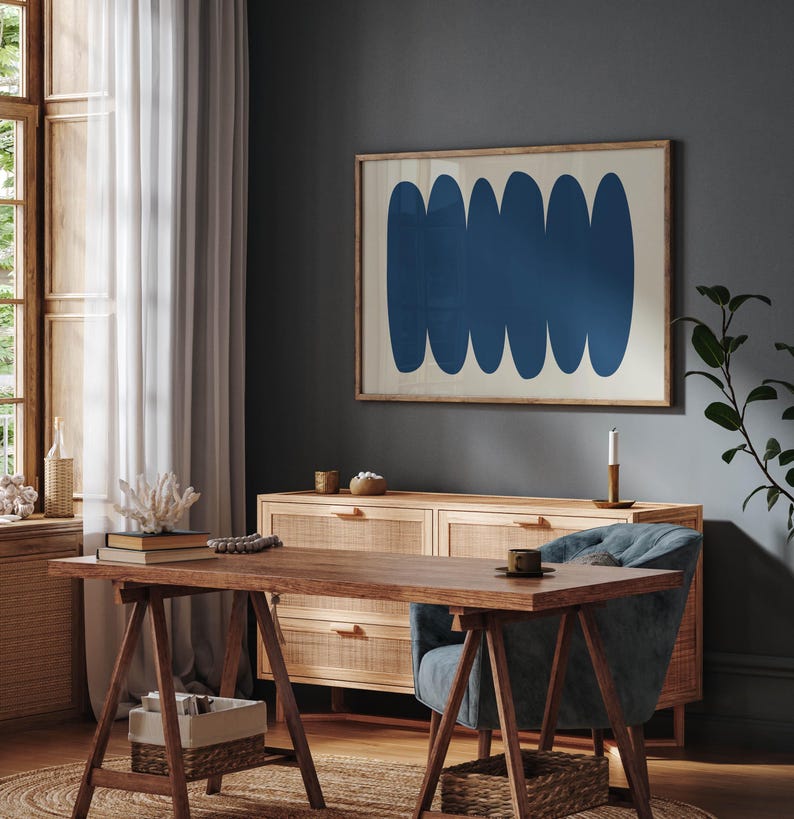 May include: A dark blue abstract art print with a white background hangs on a dark gray wall above a wooden dresser with woven cane drawers. A blue armchair sits in front of the dresser, and a wooden table with a trestle base sits in front of the chair.