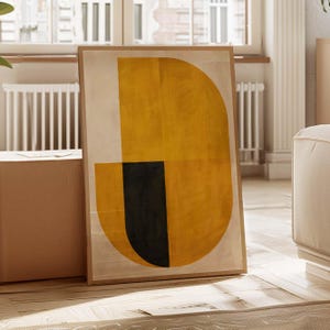 May include: Framed abstract art print with a geometric design. The artwork features a large yellow semi-circle and a black vertical rectangle against a beige background. The framed art is leaning against a wall in a room with natural light.