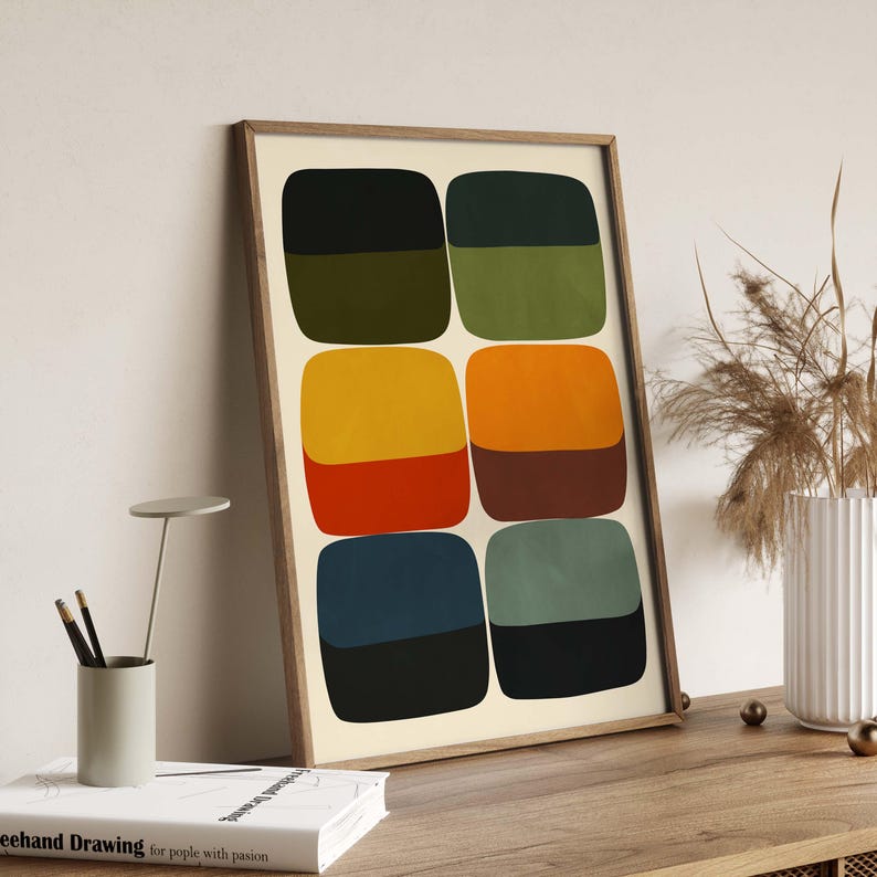 May include: A framed abstract art print featuring six pairs of rounded squares in various colors: olive green, yellow, orange, red, navy, and teal. The top halves are darker shades, while the bottom halves are lighter. The artwork is displayed on a wooden surface.