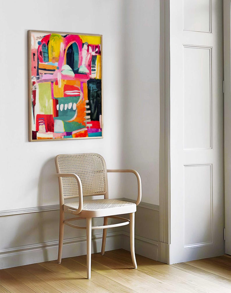 May include: A light wood chair with a woven cane seat and back sits in a corner of a room with white walls and a framed abstract painting with bright colours, including pink, yellow, green, and blue.