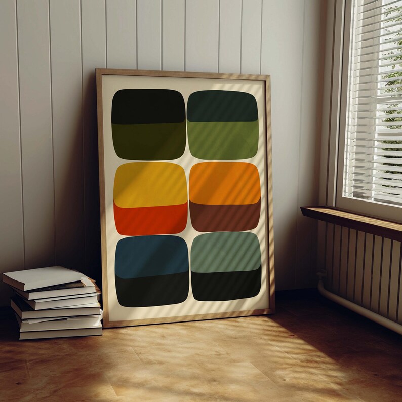 May include: A framed abstract art print featuring six pairs of rounded rectangles in various colors: olive green, yellow, orange, navy blue, and black. The artwork is displayed against a neutral background, leaning against a wall near a window.