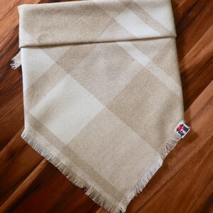 May include: A beige and white plaid dog bandana with a frayed edge. The bandana has a small logo on the bottom right corner.