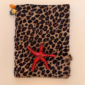 Puede incluir: Una funda rectangular para cuaderno con un estampado de leopardo en marrón y negro y una textura similar al pelaje. Un dije de estrella de mar de color rojo brillante está sujeto a una banda elástica negra y dos pequeñas cuentas en la esquina superior izquierda.