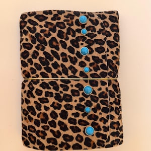 May include: A rectangular notebook covered in a brown and black leopard print fur-like material. The front flap is secured by a thin gold-coloured elastic band and is decorated with seven round, bright blue turquoise-coloured cabochon studs.