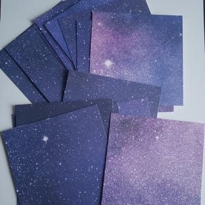 May include: A collection of square paper sheets with a starry night design. The paper features a gradient of dark blue to purple, speckled with white stars and a bright, central star. The sheets are arranged in a layered pattern.