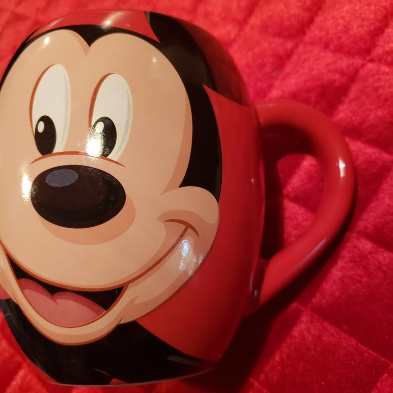 May include: A red ceramic mug featuring a smiling cartoon face with black, white, and pink details. The mug has a red handle and a glossy finish, perfect for hot beverages.