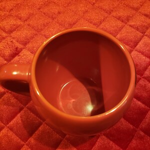 May include: A top-down view of a large, reddish-orange ceramic mug with a rounded handle. The mug is empty and sits on a textured, quilted, reddish-orange surface. The mug's interior is smooth and glossy.