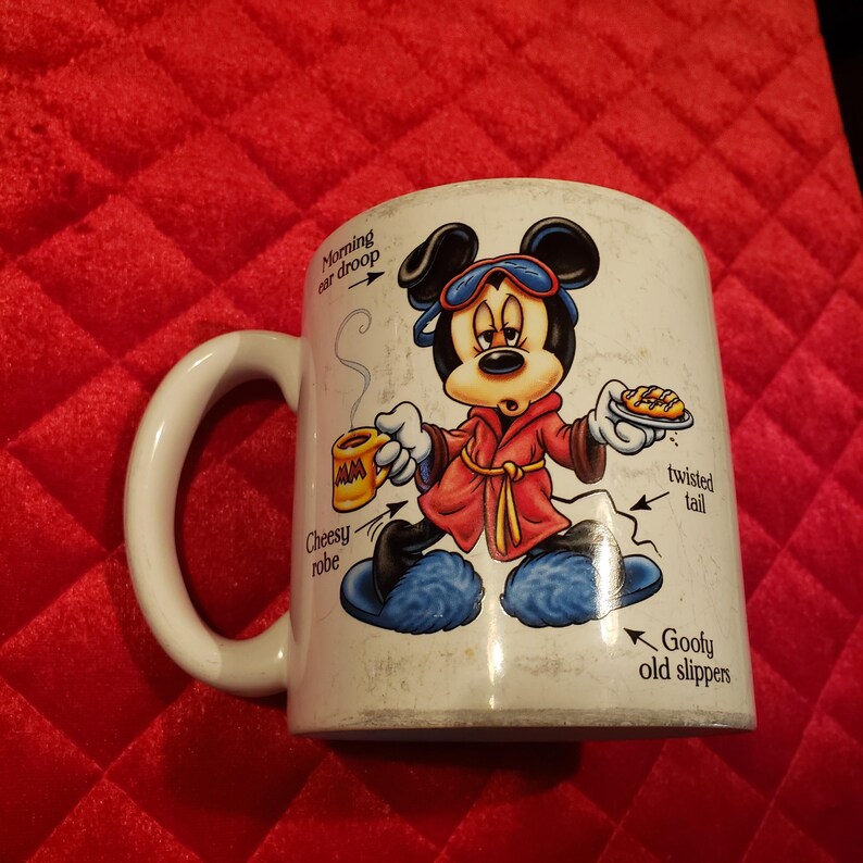 Puede incluir: Taza de cer&aacute;mica blanca con Mickey Mouse de dibujos animados con una bata roja y zapatillas azules. Mickey sostiene una taza de caf&eacute; y un plato de comida. La taza tiene un asa blanca y texto: "Morning ear droop", "Cheesy robe", "twisted tail" y "Goofy old slippers".