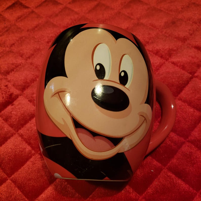 May include: A red ceramic mug featuring a smiling Mickey Mouse face. The mug has a red handle and a black and red design on the sides. The background is a red, textured surface.