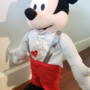 K&ouml;nnte beinhalten: Eine Pl&uuml;schpuppe von Mickey Mouse mit einem wei&szlig;en Hemd, roten Shorts und schwarzen Schuhen. Die Puppe hat eine wei&szlig;-graue Krawatte mit Zickzackmuster, graue Hosentr&auml;ger und ein rotes Herzdetail. Die Ohren der Puppe sind schwarz.