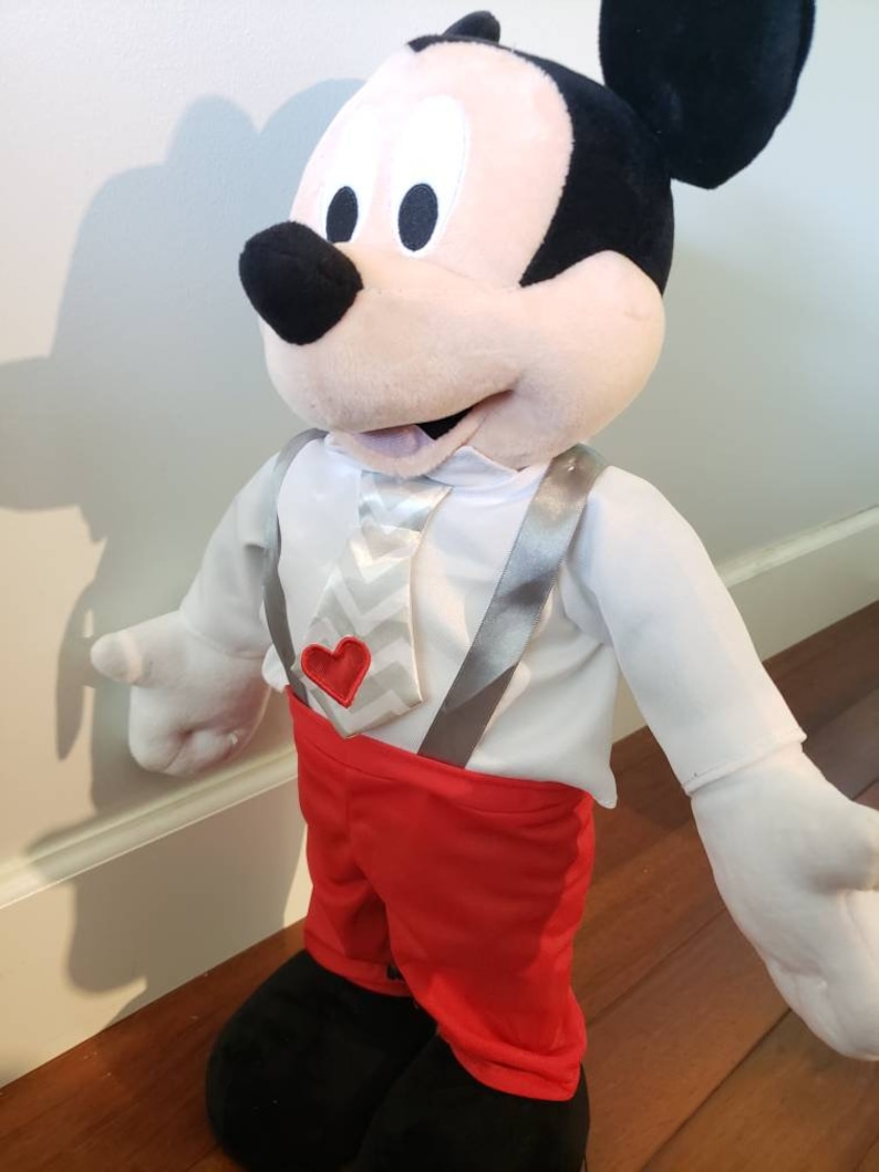 K&ouml;nnte beinhalten: Eine Pl&uuml;schpuppe von Mickey Mouse mit einem wei&szlig;en Hemd, roten Shorts und schwarzen Schuhen. Die Puppe hat eine wei&szlig;-graue Krawatte mit Zickzackmuster, graue Hosentr&auml;ger und ein rotes Herzdetail. Die Ohren der Puppe sind schwarz.