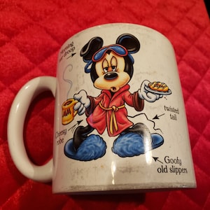 Puede incluir: Taza de cer&aacute;mica blanca con Mickey Mouse con bata roja, zapatillas azules y antifaz. La taza tiene un asa blanca e ilustraciones con texto que incluye "Morning eye droop", "Cheesy robe", "Twisted tail" y "Goofy old slippers".
