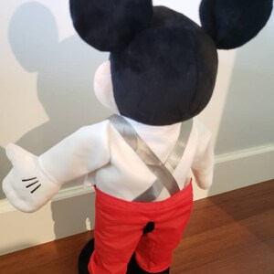 K&ouml;nnte beinhalten: Eine Mickey Mouse-Pl&uuml;schpuppe, von hinten gesehen, mit schwarzen Ohren, einem wei&szlig;en Hemd, einer roten Hose und schwarzen Schuhen. Die Puppe hat eine wei&szlig;e behandschuhte Hand mit drei schwarzen Linien. Die Hosentr&auml;ger der Puppe sind silberfarben.