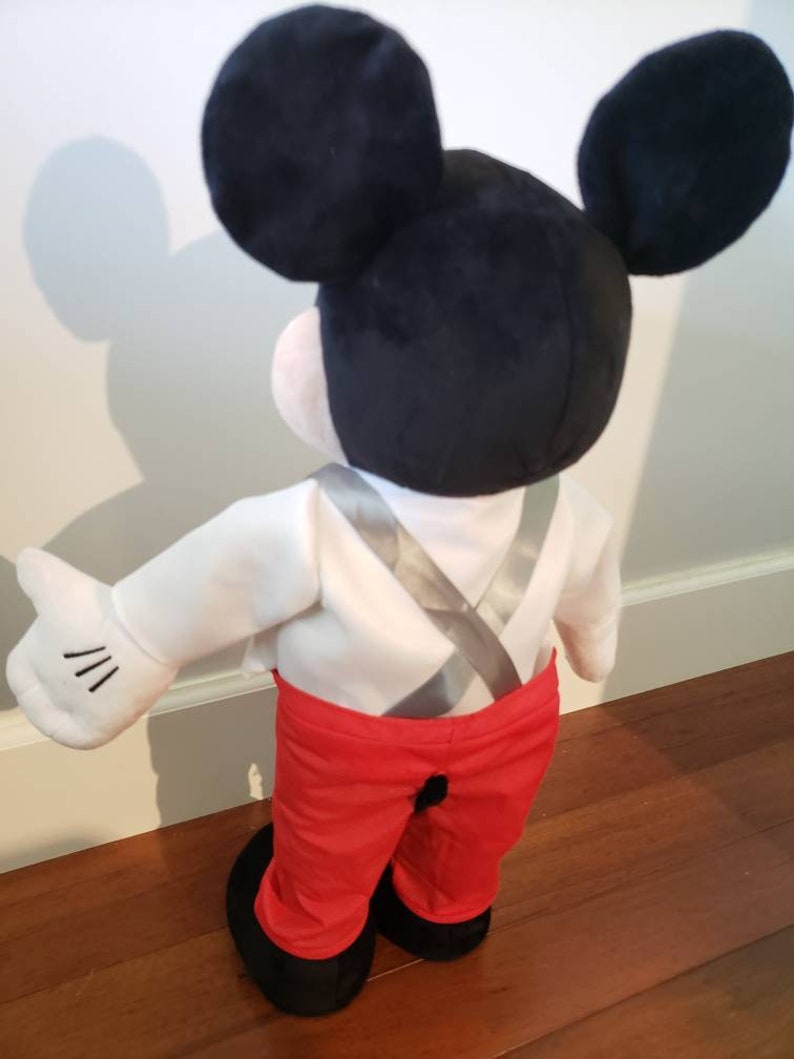 K&ouml;nnte beinhalten: Eine Mickey Mouse-Pl&uuml;schpuppe, von hinten gesehen, mit schwarzen Ohren, einem wei&szlig;en Hemd, einer roten Hose und schwarzen Schuhen. Die Puppe hat eine wei&szlig;e behandschuhte Hand mit drei schwarzen Linien. Die Hosentr&auml;ger der Puppe sind silberfarben.
