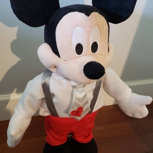 K&ouml;nnte beinhalten: Pl&uuml;schtier Mickey Mouse mit schwarzen Ohren, Nase und wei&szlig;em Gesicht. Mickey tr&auml;gt ein wei&szlig;es Hemd mit grauen Hosentr&auml;gern, rote Shorts und schwarze Schuhe. Ein herzf&ouml;rmiges rotes Emblem befindet sich auf dem Hemd.
