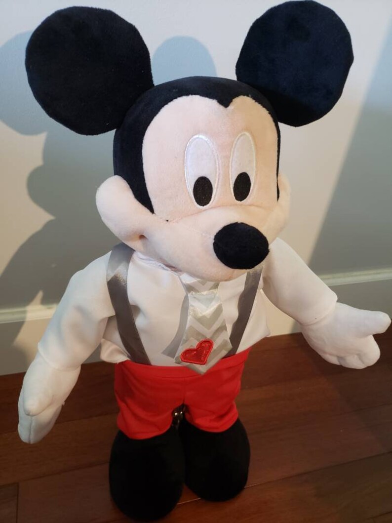 K&ouml;nnte beinhalten: Pl&uuml;schtier Mickey Mouse mit schwarzen Ohren, Nase und wei&szlig;em Gesicht. Mickey tr&auml;gt ein wei&szlig;es Hemd mit grauen Hosentr&auml;gern, rote Shorts und schwarze Schuhe. Ein herzf&ouml;rmiges rotes Emblem befindet sich auf dem Hemd.