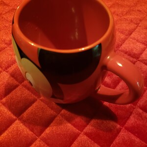 May include: Red ceramic mug featuring a Mickey Mouse design. The mug has a rounded shape with a large handle. The Mickey Mouse design includes black and white elements. The mug is sitting on a red, quilted surface.