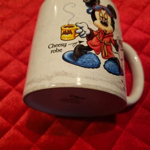 Puede incluir: Taza de cer&aacute;mica blanca con Mickey Mouse con una bata roja, sosteniendo una taza amarilla. La taza tiene el texto "Cheesy robe" y un patr&oacute;n similar al m&aacute;rmol. Tiene un asa blanca y el logotipo de Disney.