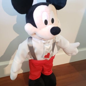 K&ouml;nnte beinhalten: Eine Mickey Mouse Pl&uuml;schpuppe mit schwarzem Kopf und gro&szlig;en Ohren, wei&szlig;em Gesicht und schwarzer Nase. Die Puppe tr&auml;gt ein wei&szlig;es Hemd, eine rote Hose und schwarze Schuhe. Sie hat graue Hosentr&auml;ger und eine Krawatte mit rotem Herz.