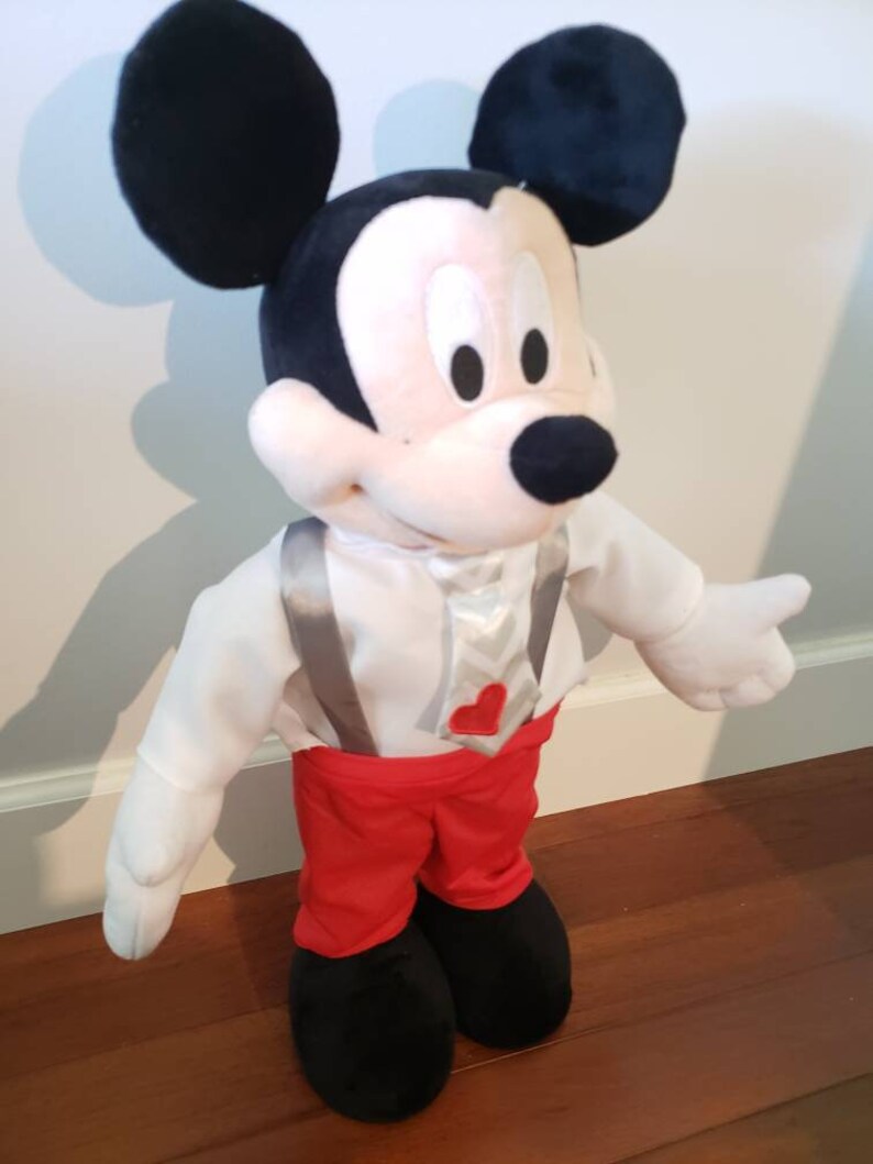 K&ouml;nnte beinhalten: Eine Mickey Mouse Pl&uuml;schpuppe mit schwarzem Kopf und gro&szlig;en Ohren, wei&szlig;em Gesicht und schwarzer Nase. Die Puppe tr&auml;gt ein wei&szlig;es Hemd, eine rote Hose und schwarze Schuhe. Sie hat graue Hosentr&auml;ger und eine Krawatte mit rotem Herz.