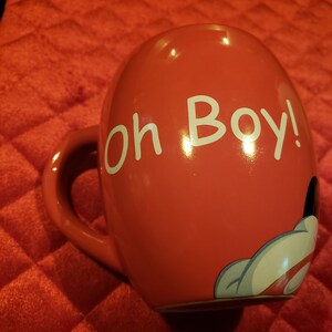 May include: A red ceramic mug with the phrase "Oh Boy!" in white lettering. The mug features a cartoon hand design in white, black, and red. The mug has a curved handle.