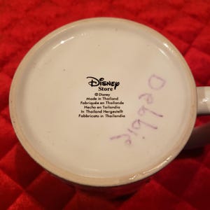 Puede incluir: Base de taza de cer&aacute;mica blanca con el logotipo de Disney Store y texto en varios idiomas. El texto incluye "Hecho en Tailandia". La palabra "Debbie" est&aacute; escrita con un rotulador morado.